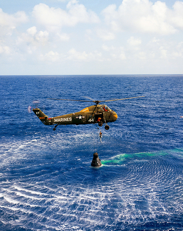 Helicopter flying over ocean winches up the Mercury-Redstone capsule which had just landed from space