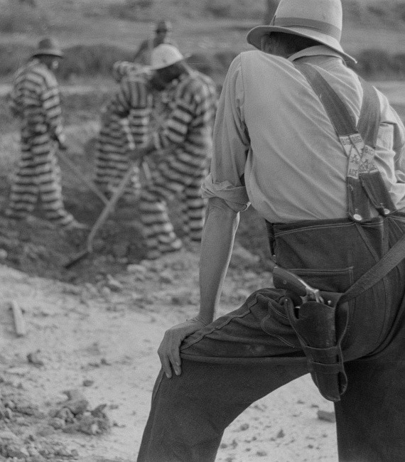 A group of incarcerated laborers dig with shovels as an armed white guard looks on